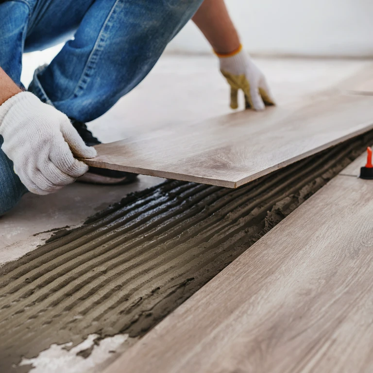 Zbliżenie na&nbsp;ręce fachowca w&nbsp;rękawicach, który kładzie panele podłogowe na&nbsp;świeżo rozprowadzoną zaprawę
