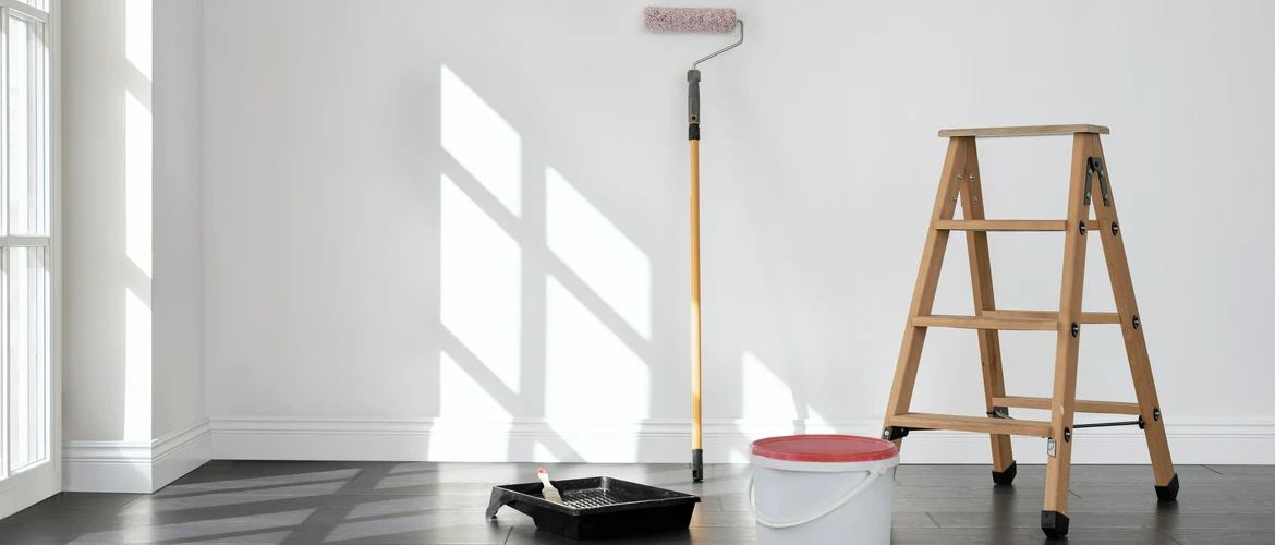 Jasne, puste pomieszczenie z&nbsp;drewnianą drabiną, wałkiem do&nbsp;malowania na&nbsp;długim kiju oraz&nbsp;wiaderkiem farby